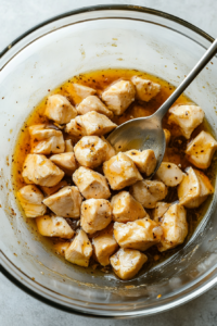 Overhead shot of a glass bowl with bite-sized chicken cubes marinating in oil, cornstarch, Shaoxing wine, salt, and white pepper. A spoon stirs the mixture, ensuring even coating.