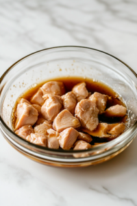 A glass bowl on a white marble cooktop containing thinly sliced chicken breast marinating in Shaoxing wine, soy sauce, and cornstarch. The chicken glistens as the marinade evenly coats each piece for a rich flavor.