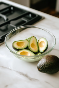 A close-up shot of a glass bowl on a white marble cooktop, where a ripe avocado is being mashed into a creamy consistency with a fork. Next to it, freshly diced avocado pieces sit, ready to be mixed in for added texture.