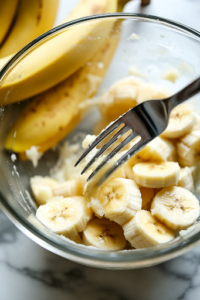 A close-up of a glass mixing bowl on a white marble cooktop, where a fork is mashing overripe bananas into a thick, slightly lumpy puree. The bananas are soft and fragrant, releasing their natural sweetness for the banana bread batter.