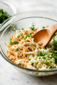 A glass mixing bowl on a white marble cooktop is filled with shredded chicken, chopped cabbage, and sliced green onions. A wooden spoon rests inside, ready to mix the fresh and vibrant ingredients.