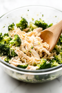 A glass bowl on a white marble cooktop filled with shredded chicken, chopped steamed broccoli, garlic powder, salt, pepper, and shredded mozzarella cheese. A wooden spoon thoroughly mixes the ingredients into a flavorful filling.