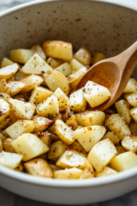 A close-up shot of a large mixing bowl filled with diced Yukon Gold potatoes, drizzled with olive oil and generously sprinkled with sea salt, black pepper, and paprika. A wooden spoon is seen tossing the potatoes, ensuring an even coating of seasoning for maximum flavor.