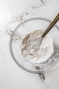 A close-up shot of a glass mixing bowl placed on a white marble cooktop, filled with whisked all-purpose flour, salt, baking powder, and baking soda. A metal whisk rests inside, mid-motion, ensuring all dry ingredients are evenly blended for the perfect pound cake texture.