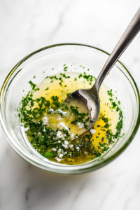 A close-up shot of a small glass bowl on a white marble cooktop, filled with softened butter, chopped parsley, minced garlic, and salt, being mixed with a spoon. The creamy mixture is well combined and ready to be frozen for later use.