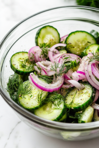 A close-up view of a glass mixing bowl on a white marble cooktop, filled with freshly sliced cucumbers, thinly sliced red onion, diced avocado, and finely chopped fresh dill. The vibrant green and purple hues of the ingredients create a visually appealing contrast.