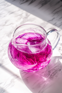 A close-up shot of a clear glass pitcher placed on a white marble cooktop, filled with freshly poured lemonade and deep purple grape juice. The two vibrant colors swirl together, creating a mesmerizing and magical base for the Potion Punch.