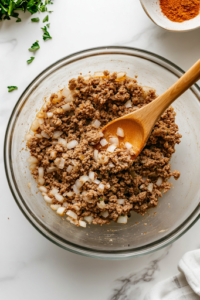A close-up shot of a large glass mixing bowl on the white marble cooktop, filled with lean ground beef, plain breadcrumbs, diced yellow onion, eggs, Worcestershire sauce, and seasonings. A wooden spoon rests on the side, ready to mix the ingredients into a well-seasoned meatloaf base.