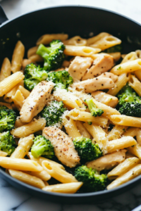 A black skillet on a white marble cooktop, now filled with cooked penne pasta, tender broccoli, and sliced chicken. Everything is being tossed together, evenly coated in the rich and velvety garlic Parmesan sauce.