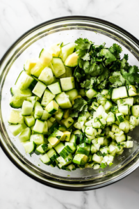 A close-up view of a glass mixing bowl on a white marble cooktop, filled with cubed cucumbers, avocados, freshly chopped cilantro, and minced garlic. The vibrant green colors of the ingredients create a refreshing and flavorful mix.