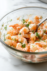 A glass mixing bowl on the white marble cooktop filled with chopped shrimp, mayonnaise, minced garlic, finely chopped parsley, and dill. A spoon is stirring the ingredients together, creating a creamy and flavorful filling.