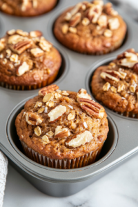 A close-up of muffins cooling on a wire rack over a white marble cooktop, with one muffin cut in half to reveal its moist, fluffy interior. A few scattered pecans and dried cherries add texture, making the muffins look even more delicious.