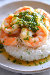 A beautifully plated dish of shrimp with salsa verde over a bed of rice, garnished with freshly chopped cilantro and a lime wedge. A refreshing cucumber salad sits in the background, completing the meal.