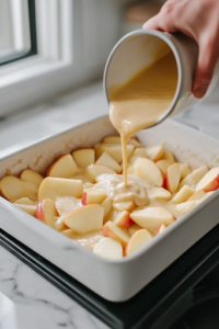 The same baking dish on the white marble cooktop as a hand pours the smooth batter evenly over the apple slices, ensuring they are fully covered and ready for baking.
