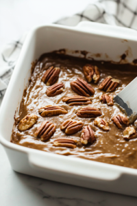 A close-up shot of a 9×13-inch baking dish placed on the white marble cooktop, with the thick pecan batter being poured in. A spatula spreads the batter evenly across the dish, ensuring a smooth and level surface before baking.