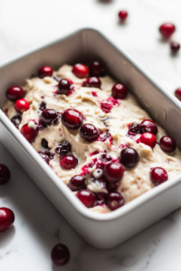 A close-up shot of the prepared loaf batter being poured into a parchment-lined loaf pan on the white marble cooktop. The cranberries are evenly distributed throughout the batter, creating a vibrant contrast in the smooth, creamy mixture.
