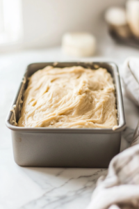 A close-up of the smooth banana bread batter being poured into a greased 9×5-inch loaf pan on a white marble cooktop. The batter spreads evenly, filling the pan and preparing to rise into a fluffy, golden loaf.