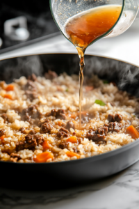 A close-up shot of a measuring cup pouring beef broth and water into the black skillet on the white marble cooktop. The liquid bubbles as it mixes with the seasoned rice and meat, preparing for slow absorption.