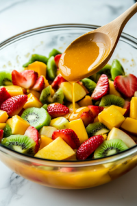 A close-up shot of the citrus dressing being poured from the small glass bowl into the large fruit-filled mixing bowl on the white marble cooktop. A wooden spoon is positioned to gently toss the salad, ensuring all the fresh fruits are evenly coated with the zesty dressing.