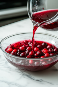 A close-up shot of deep red cranberry juice being poured into the glass punch bowl on the white marble cooktop. The rich liquid flows over the fresh fruit, blending into the bowl and creating a flavorful and colorful base for the Christmas Punch.