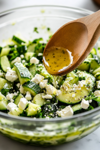 A close-up view of a glass bowl on a white marble cooktop, where the freshly prepared dressing is being poured over a mixture of cucumbers, avocado, and feta. A wooden spoon is positioned nearby, ready to toss the salad evenly.