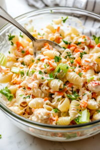 A large spoon is stirring the crab pasta salad, ensuring all ingredients are evenly coated with the rich and creamy dressing. The glass mixing bowl sits on the white marble cooktop, with the salad looking vibrant and well-mixed.