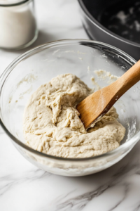 A close-up view of a glass mixing bowl on a white marble cooktop as hot water is gradually poured in. A wooden spoon stirs the mixture, forming a soft and pliable tortilla dough.