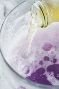 A close-up shot of a cold bottle of lemon-lime soda being carefully poured into the glass pitcher on the white marble cooktop. Tiny bubbles rise to the surface, adding an effervescent sparkle and a refreshing fizz to the Potion Punch.