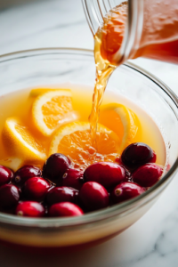 A close-up shot of golden orange juice being poured into the punch bowl on the white marble cooktop, mixing with the cranberry juice to create a beautiful layered effect. The fresh citrus aroma enhances the festive essence of the drink.