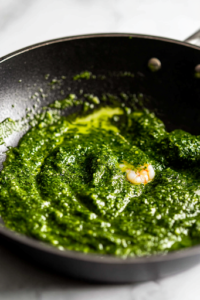 A black skillet on a white marble cooktop with vibrant green salsa verde being poured over the shrimp. The sauce coats each piece, gently simmering as the flavors meld together.