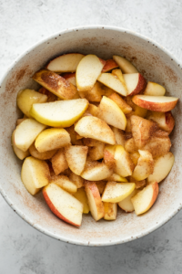 An overhead shot of a mixing bowl filled with freshly peeled, cored, and sliced Granny Smith apples. The apples are coated with white sugar, cinnamon, and nutmeg, giving them a glossy, spiced appearance, ready to be baked into the crisp.