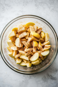 An overhead shot of a large mixing bowl filled with freshly peeled, cored, and sliced Granny Smith apples. The apples are coated in sugar, flour, lemon juice, zest, cinnamon, and nutmeg, creating a glossy, spiced mixture, ready to be used in the pie.