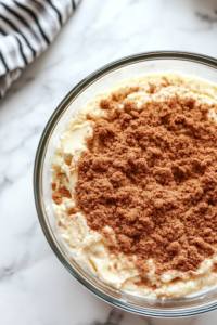A close-up shot of a second glass mixing bowl on a white marble cooktop, where softened butter, brown sugar, flour, and cinnamon are being mixed together with a fork. The mixture is thick and crumbly, forming a rich and aromatic cinnamon topping that will add a sweet and spiced swirl to the cake.
