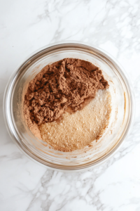 A close-up shot of another glass mixing bowl on a white marble cooktop, filled with flour, baking powder, cinnamon, baking soda, and salt. A wooden spoon is stirring the dry ingredients thoroughly to ensure an even distribution of flavors and leavening agents.