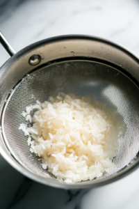 A close-up shot of a fine mesh strainer placed over the white marble cooktop, with Calrose rice being rinsed under cold running water. The water appears cloudy at first, gradually turning clear as excess starch is washed away.