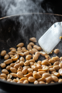 Close-up shot of raw peanuts roasting in a wok with a teaspoon of vegetable oil, being stirred with a spatula. The peanuts turn golden brown as steam rises from the pan.
