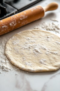 A close-up view of a wooden rolling pin flattening a dough ball into a thin, round tortilla on a floured surface over the white marble cooktop. The dough stretches evenly, preparing for cooking.