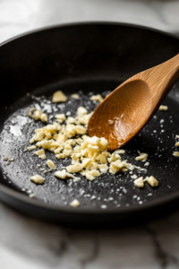 A black skillet on a white marble cooktop, where minced garlic sizzles in the remaining butter and oil. A wooden spoon gently stirs, releasing the rich, fragrant aroma as the garlic turns golden.