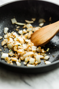 A black skillet on a white marble cooktop contains minced onions, garlic, ginger, and serrano pepper sautéing in melted ghee. The mixture turns golden and releases a rich aroma as a wooden spoon gently stirs the ingredients.