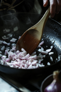 A black skillet on a white marble cooktop now containing minced shallots (or red onions) being sautéed in oil. A wooden spoon stirs the translucent aromatics, enhancing their natural sweetness.