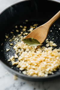 A close-up shot of a black pot on a white marble cooktop, where ghee is melting. Chopped garlic, ginger, and a single bay leaf are sizzling, releasing a rich and aromatic fragrance. A wooden spatula stirs the ingredients, ensuring even cooking