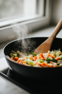 A black skillet on a white marble countertop where chopped garlic, onions, shredded carrots, and celery are being sautéed in olive oil. Steam rises as the vegetables soften, while a wooden spoon stirs the mixture to enhance their aroma and flavor.