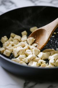 A large black pot sits on a white marble cooktop, where diced boneless skinless chicken thighs sizzle in olive oil. The chicken is turning golden brown with a light sear as a wooden spoon gently stirs the pieces, releasing a savory aroma.