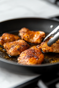 A close-up shot of a black skillet on a white marble cooktop, where seasoned chicken thighs sizzle in hot oil. The golden-brown crust is forming as tongs lift one piece, revealing a crispy sear that locks in flavor and moisture.