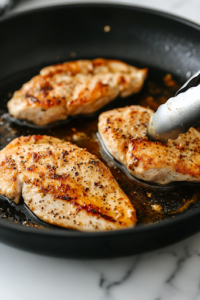 Seasoned chicken breasts sizzle in a black skillet on a white marble cooktop, forming a golden-brown crust. A pair of tongs is ready to flip one of the pieces, ensuring even cooking.