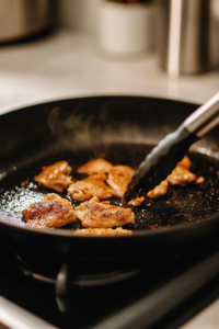 A black skillet on a white marble cooktop sizzles with hot oil as golden-brown chicken pieces cook evenly. A pair of tongs carefully flips a piece, revealing a crispy, well-seared surface.