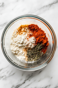 A glass bowl on the white marble cooktop holds a mixture of all-purpose flour and spices, including paprika, thyme, garlic salt, and cayenne pepper. A fork rests on the side, ready for mixing, ensuring an even distribution of flavors for coating the chicken.