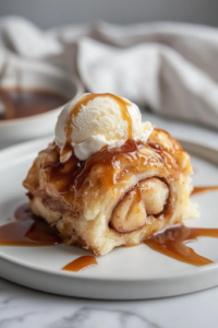 A close-up of a serving plate on a white marble cooktop, featuring a freshly baked portion of Caramel Apple Cinnamon Roll Bake. The warm dessert is drizzled with extra caramel sauce and served with a scoop of melting vanilla ice cream, ready to be enjoyed.