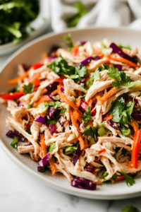 A serving plate on a white marble cooktop showcases the fully mixed Chinese Chicken Salad, bursting with crisp textures and vibrant colors. Fresh herbs garnish the dish, making it ready to be served.