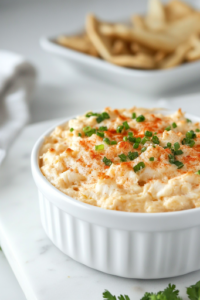 A chilled crab dip served in a white bowl on the white marble cooktop, surrounded by crispy crackers, fresh veggies, and slices of bread. The dip is creamy and ready to be enjoyed as a delicious appetizer.